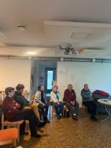 Man sieht einen halben Sitzkreis mit arbeitenden Menschen und im Hintergrund Zahlen an der Wand